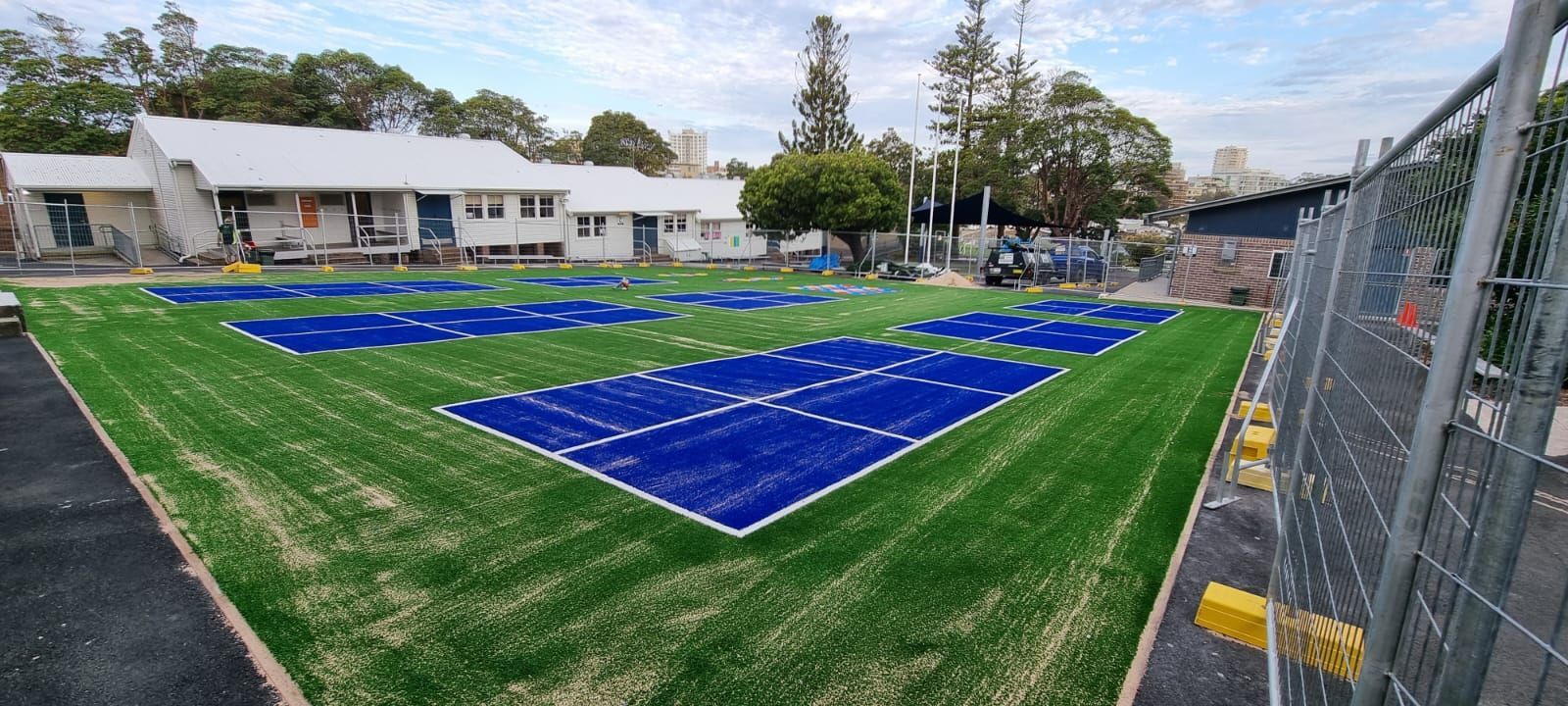 Outdoor sports field with blue courts on green turf beside a building and chain-link fence