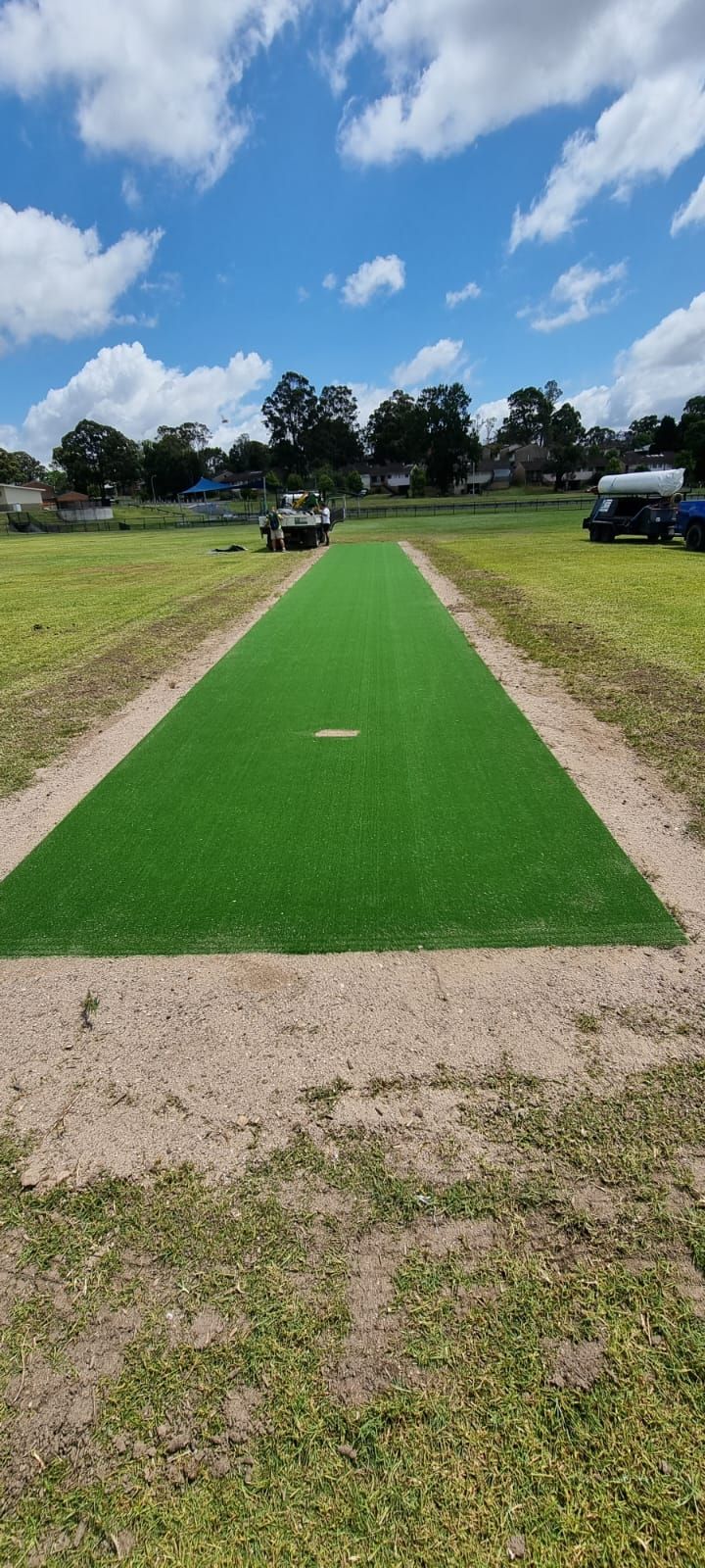 Green artificial turf runway centered on a grassy field under a blue sky with clouds