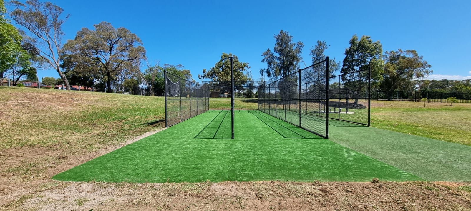 Outdoor sports court with green turf and black netting on a sunny field, surrounded by trees and grass.