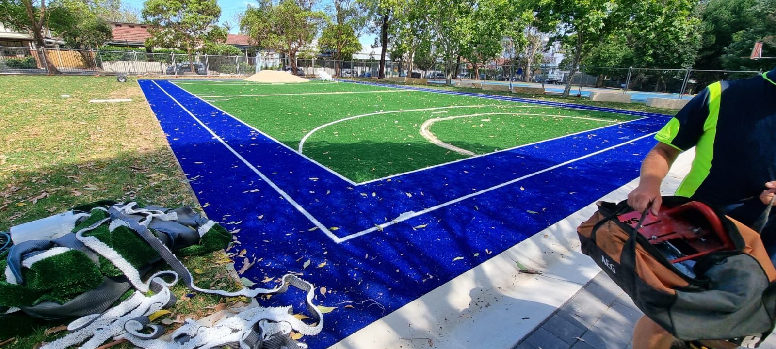 Blue-and-green sports court being freshly painted outdoors, with tools and paint cans nearby