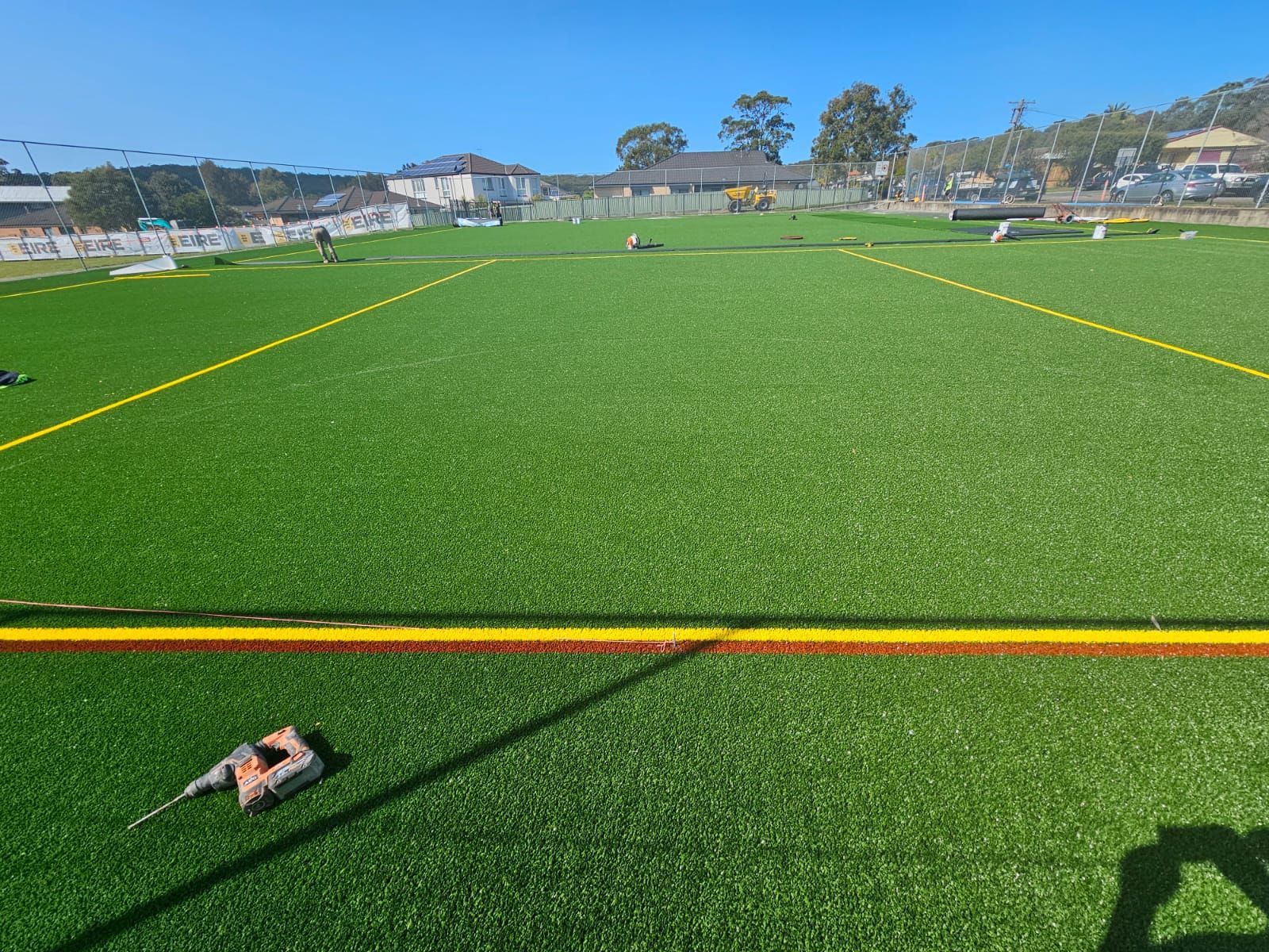 Wide green sports field with orange and yellow boundary lines, seen under a clear blue sky.