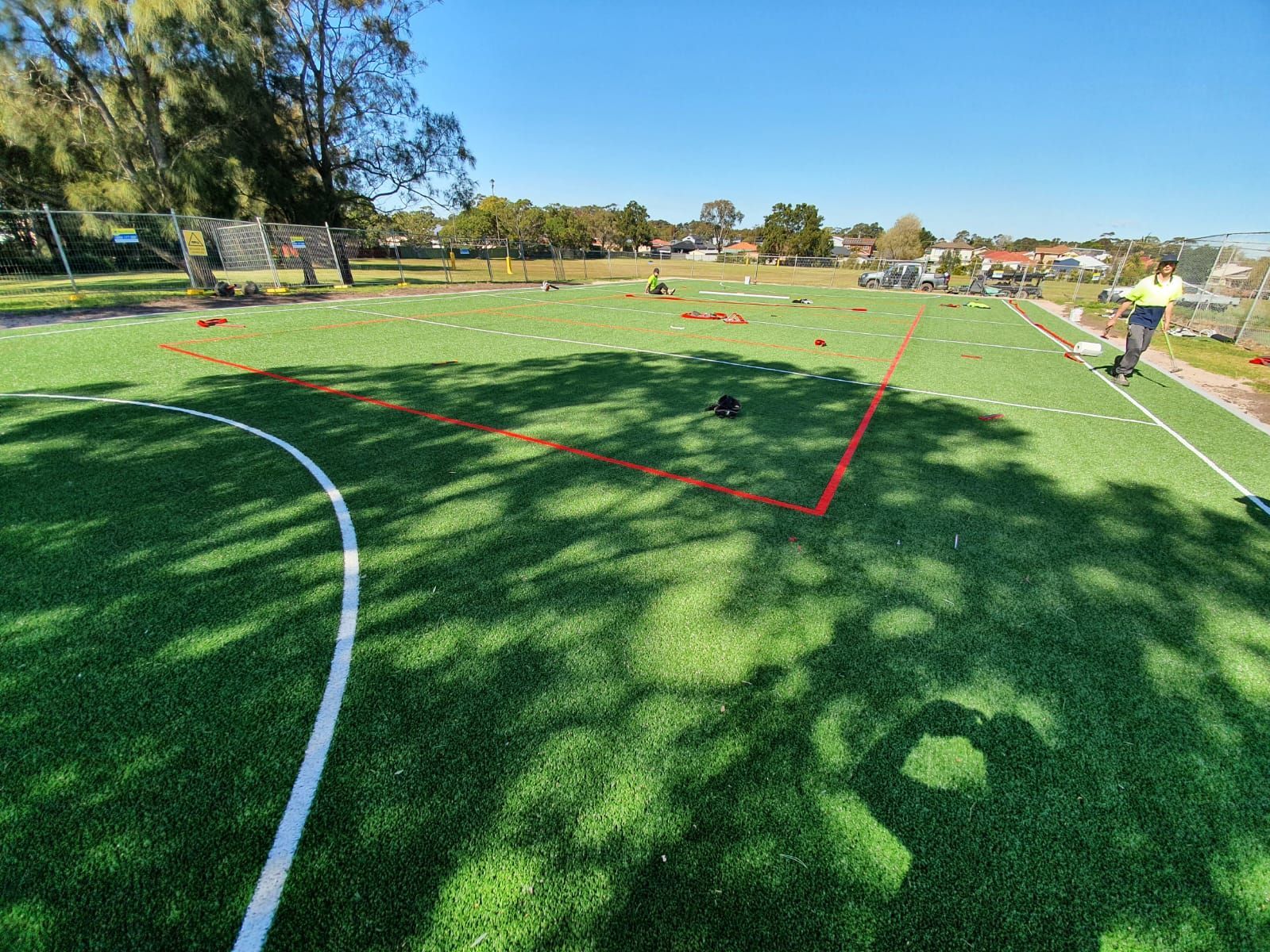 Marked sports field with white and red boundary lines, trees and houses in the background, people in the distance