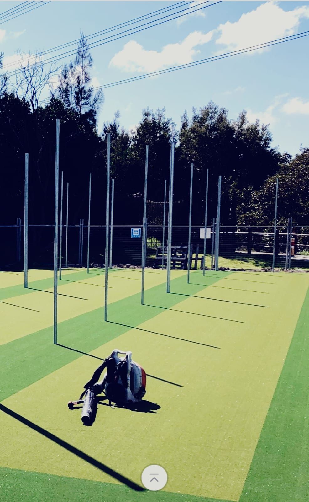 Outdoor sports field with green turf, tall poles, and a backpack in the foreground under a blue sky