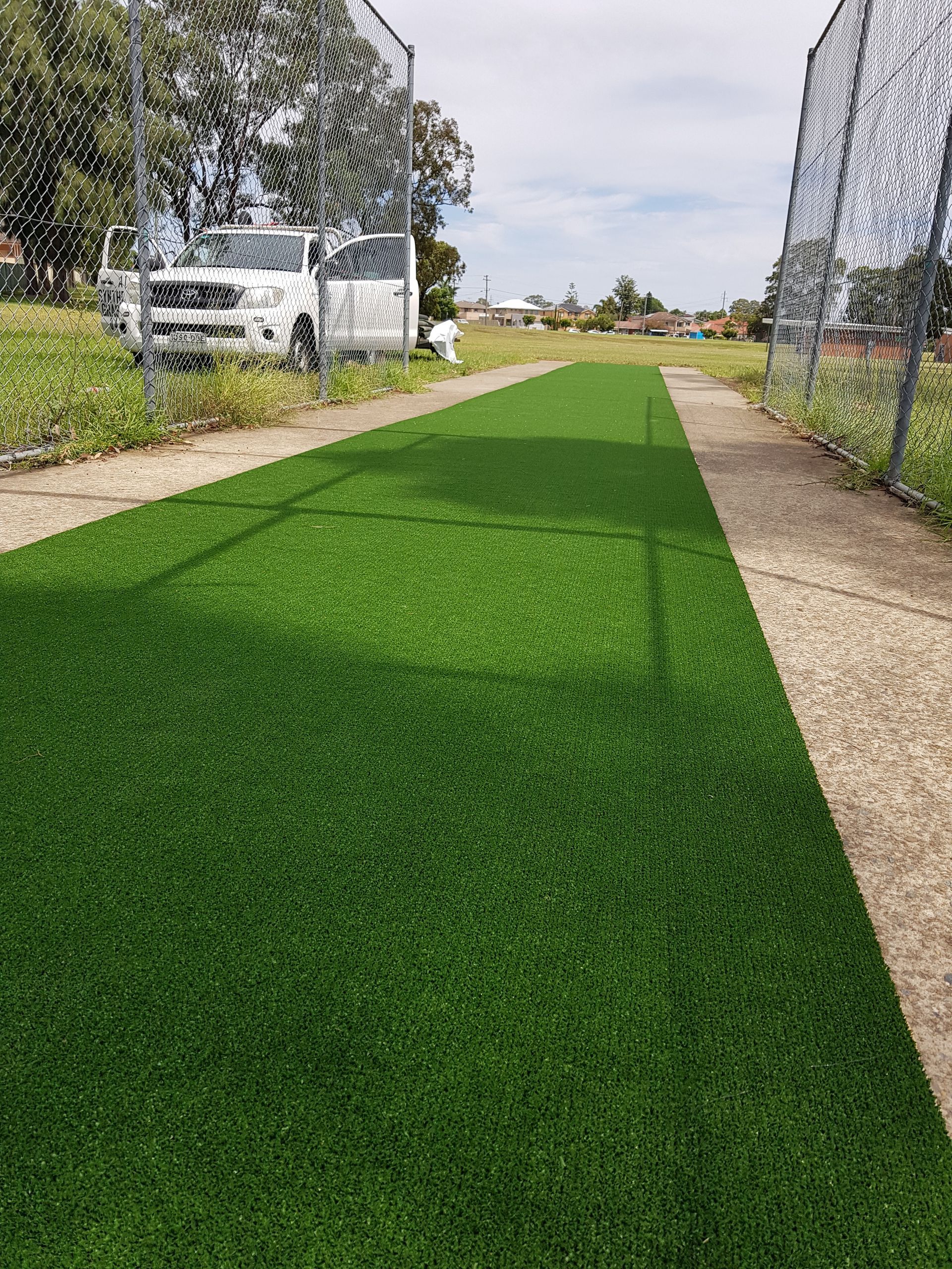 Green artificial turf lane beside a chain-link fence, with parked vehicles and trees in the background