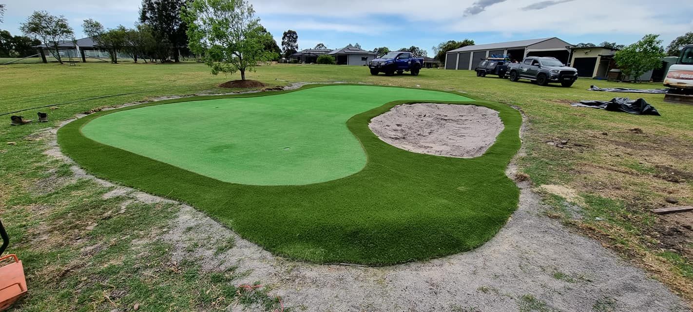 Outdoor mini-golf green with a sand bunker, surrounded by grass under a cloudy sky.