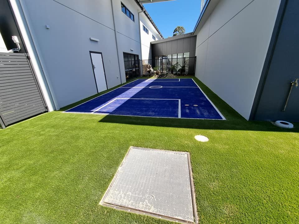 Blue backyard sports court between white buildings with green artificial turf foreground