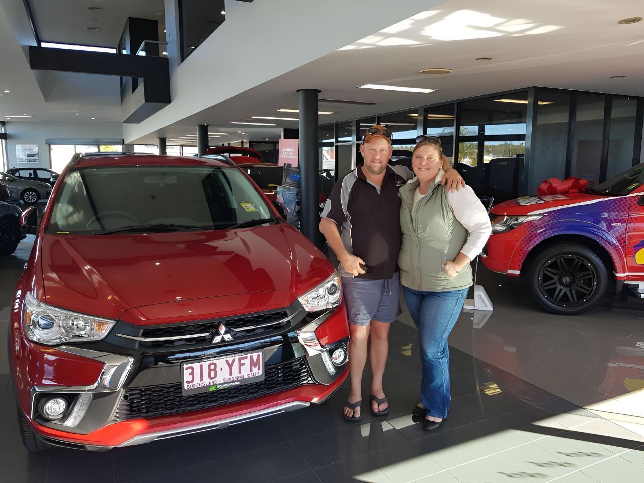 A Man And Woman Are Standing In Front Of A Red Car In A Showroom — Grant Finance Solutions In Toowoomba, QLD