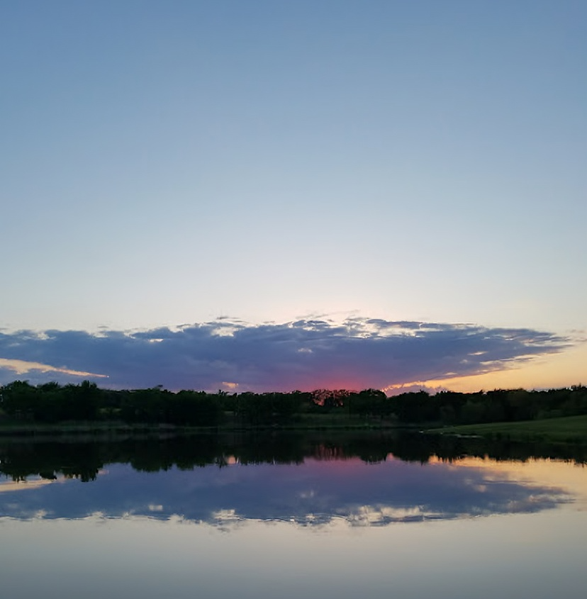 Sunset over a calm lake, reflecting sky and silhouetted trees. Pink and orange hues in clouds.