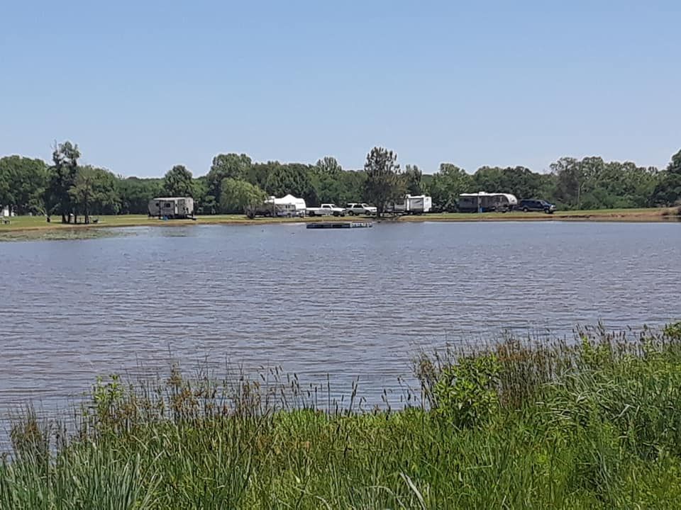Lake with RVs on the far shore, tall green grass in foreground under a bright blue sky.