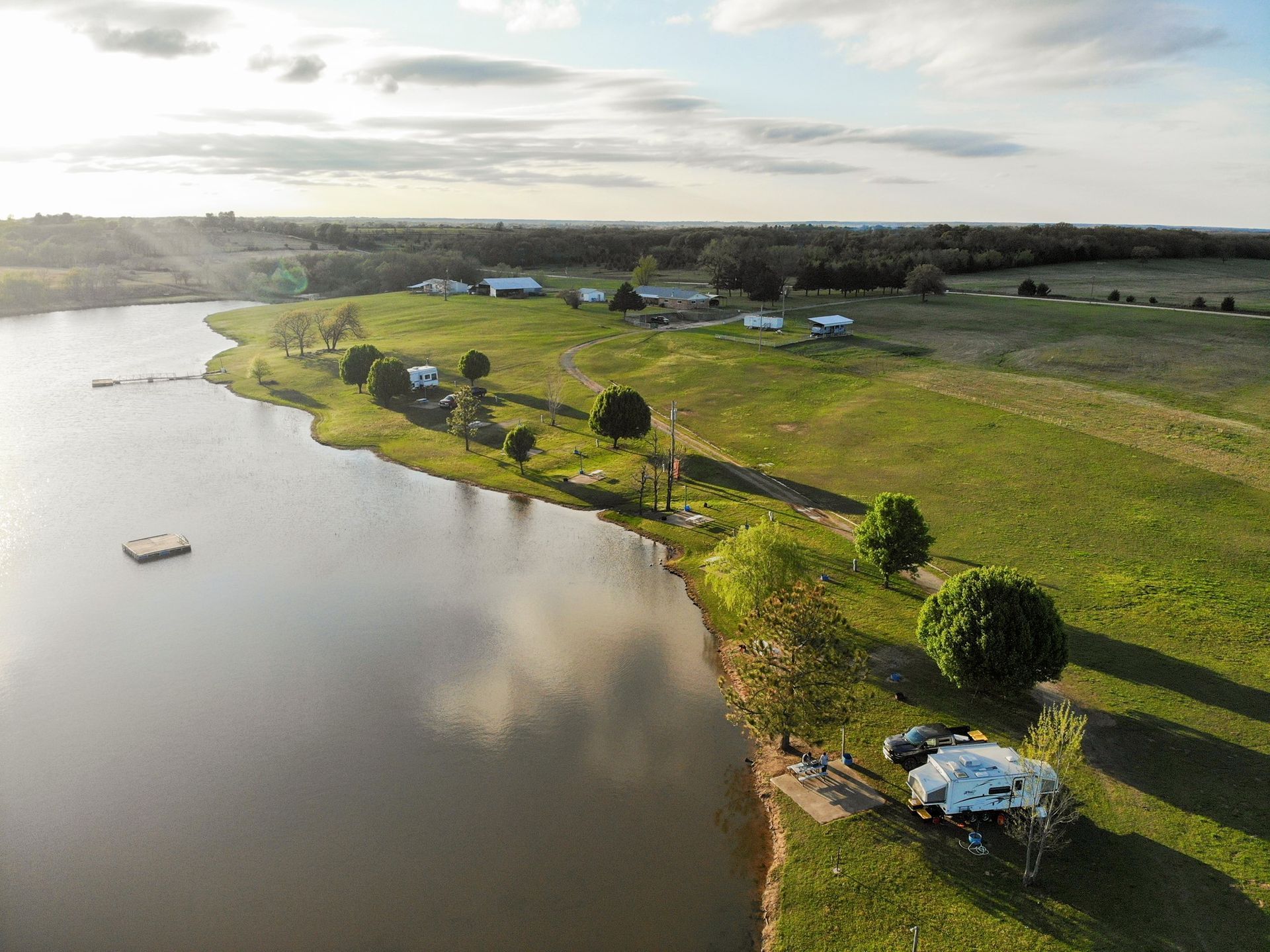 Aerial view of a lake and green pasture with buildings, a dock, and a camper on the shore under a partly cloudy sky.