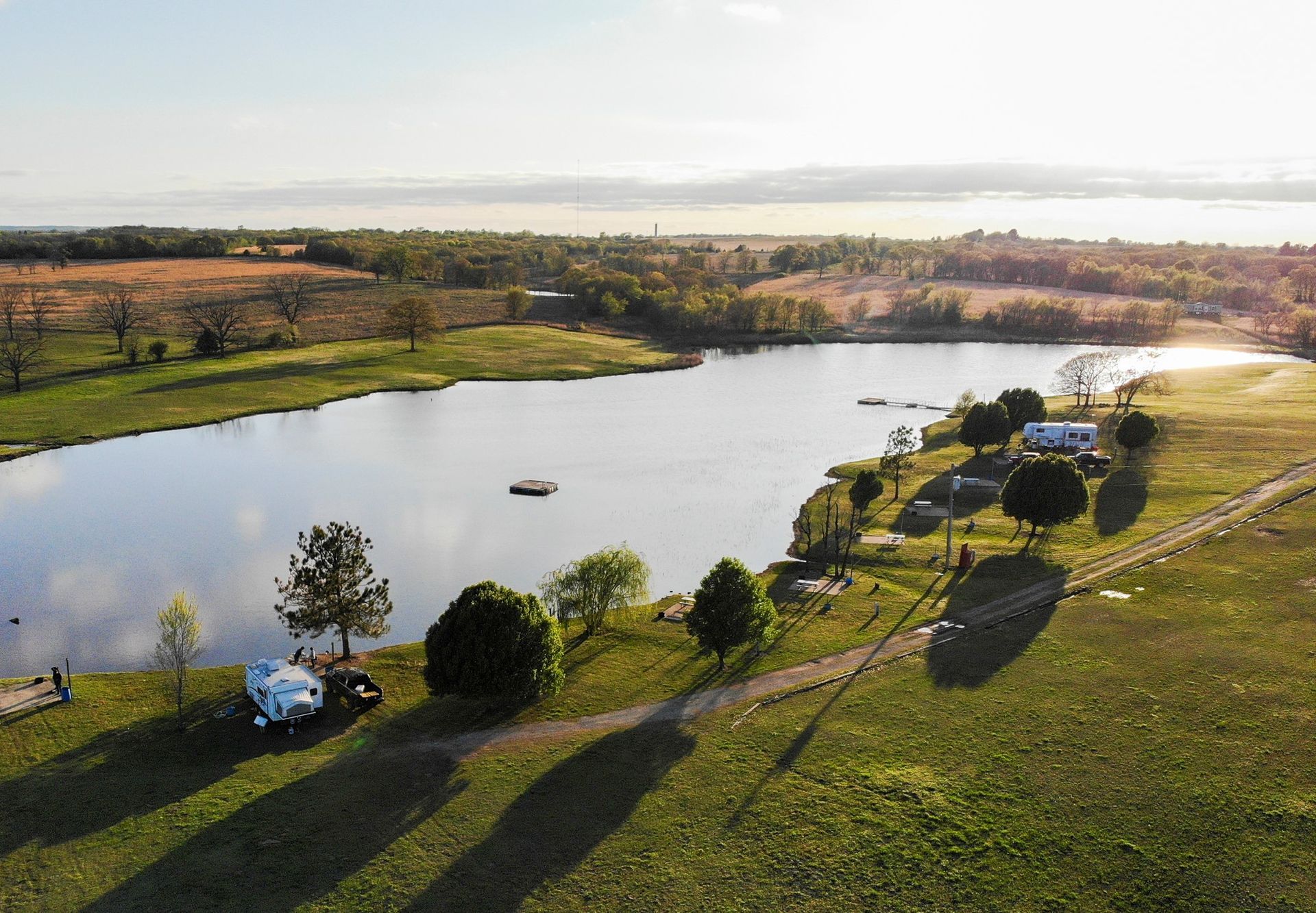 Aerial view of a lake and green grassy fields at sunset. Buildings and trees dot the shoreline.