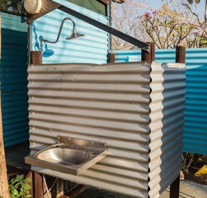 Outdoor shower stall with corrugated metal walls, a sink, and a showerhead; blue wall behind.