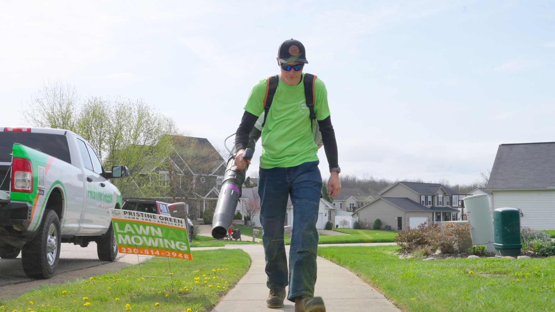 Worker using a leaf blower walks along a sidewalk beside a lawn care truck and manicured grass.