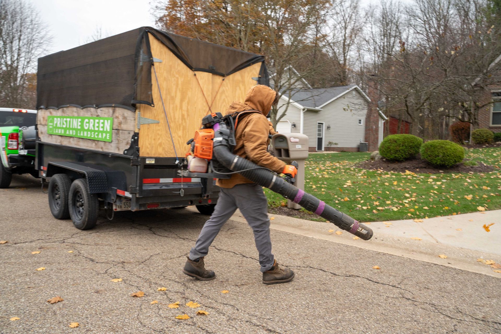 Man with leaf blower near a trailer, blowing leaves on a street. Trailer has 