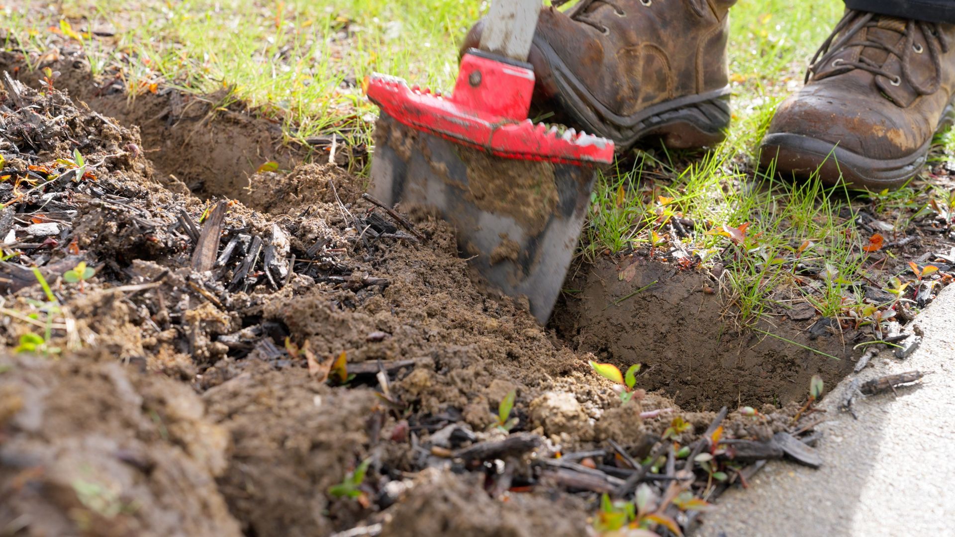 A shovel clearing dirt from a grassy edge beside a sidewalk, with boots visible.