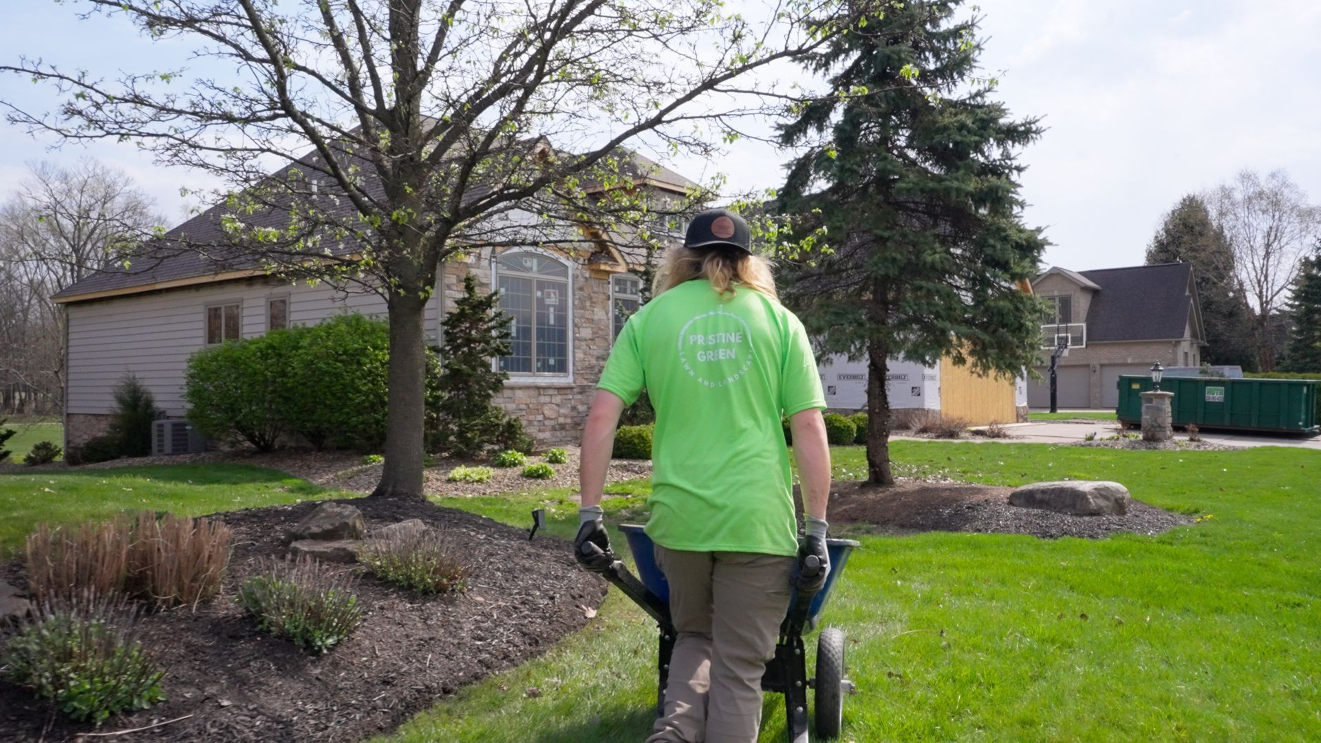 Person in bright green shirt pushing a stroller on a grassy front lawn near a suburban house.
