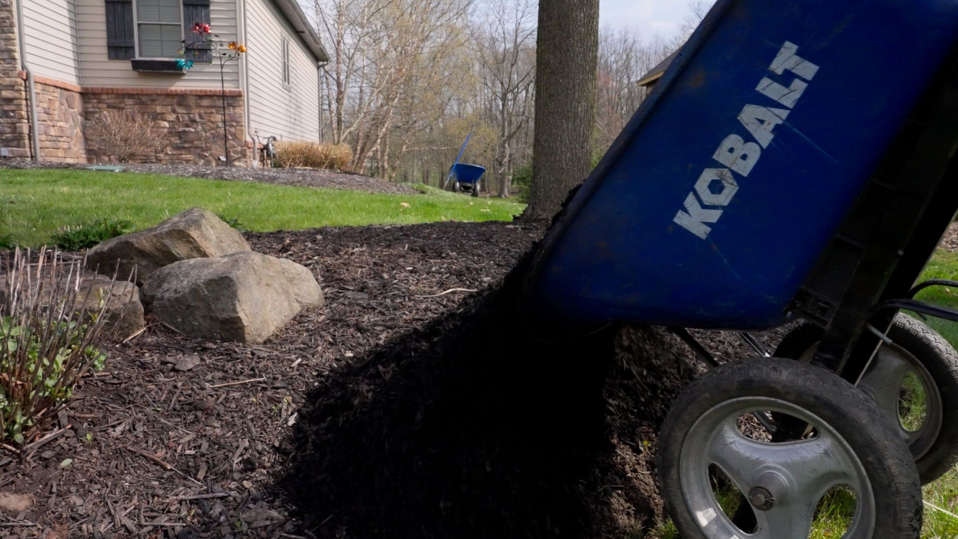 A person pours black mulch from a blue Kobalt cart beside a house and rocks in a yard.