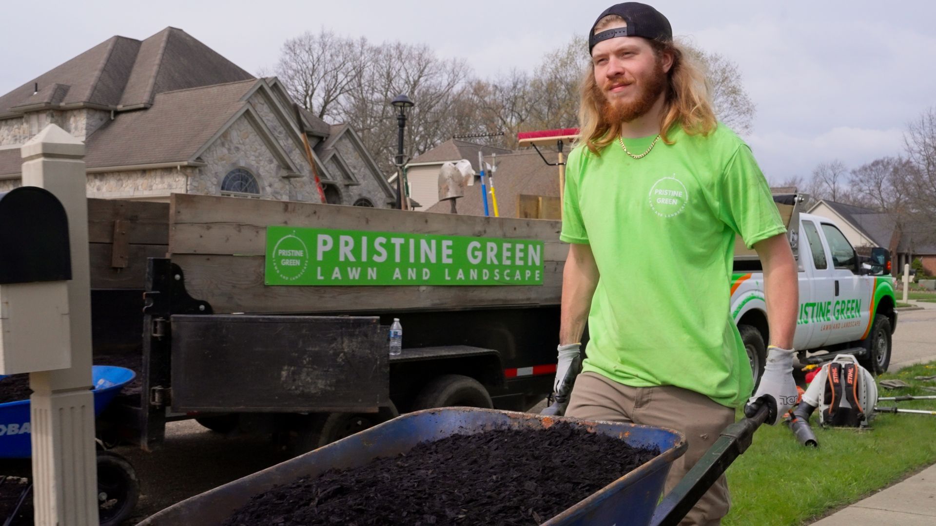 Landscaping worker in neon green shirt pushing a wheelbarrow beside a Pristine Green Lawn Care truck