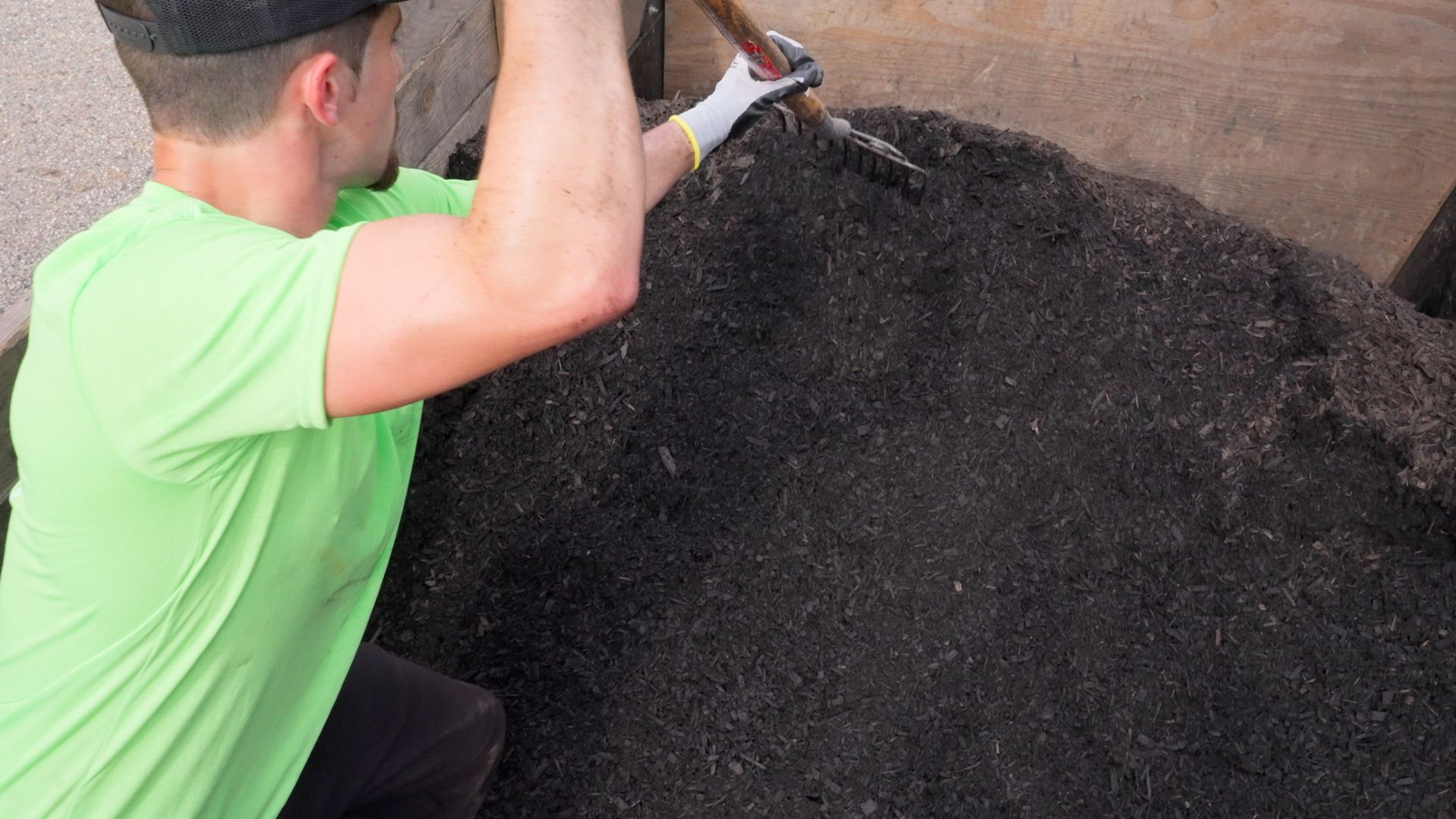 Person in a green shirt shovels dark material onto a large pile outdoors.