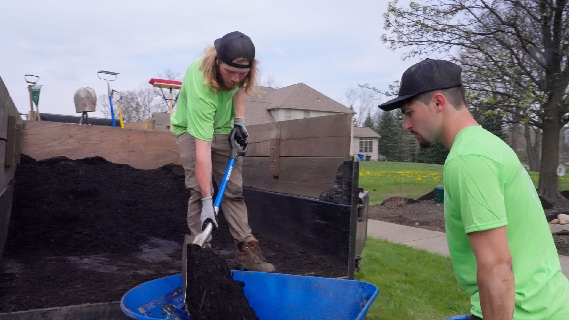 Two people in neon green shirts shoveling dirt into blue bins outdoors.