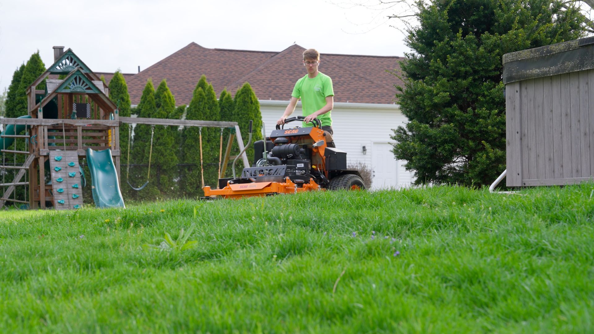 Person mowing a grassy yard on an orange riding mower near a house and trees.
