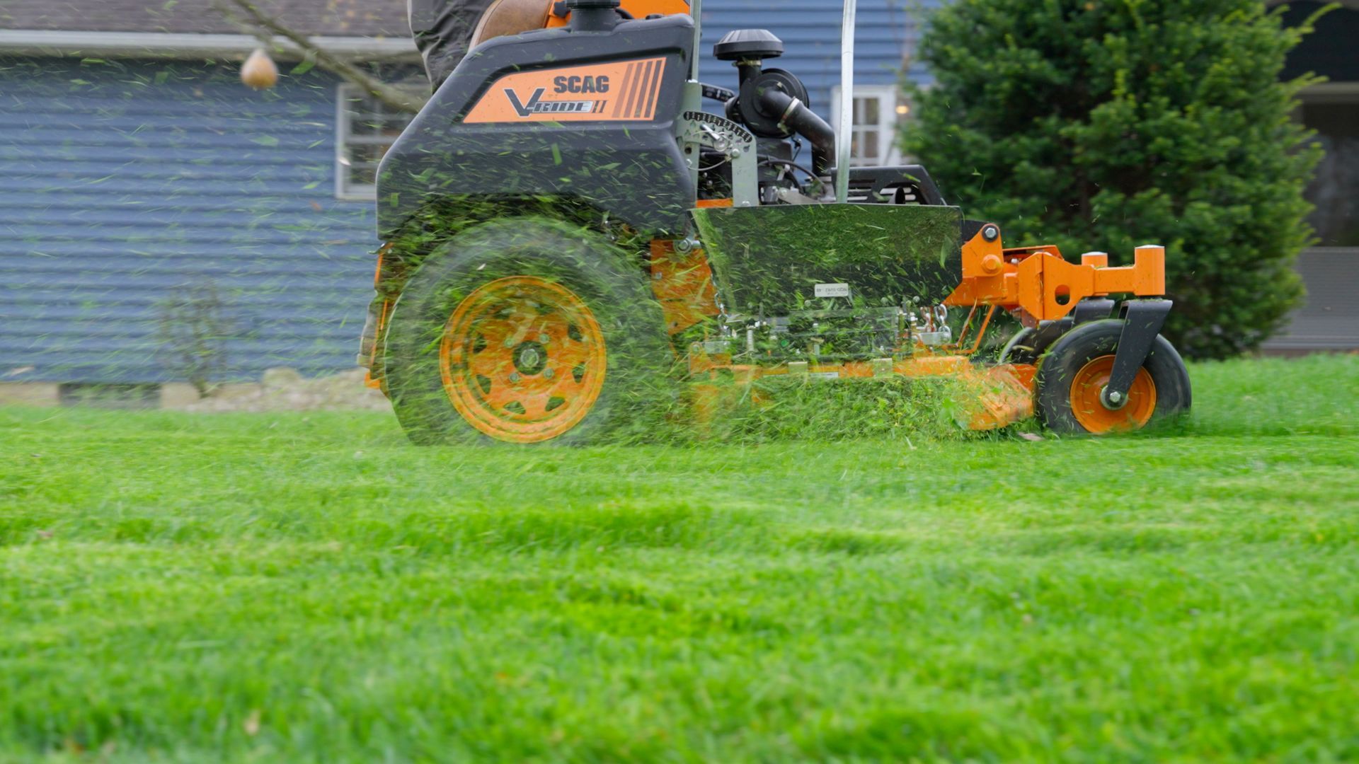 Orange lawn mower cutting grass in a green field near a house and trees