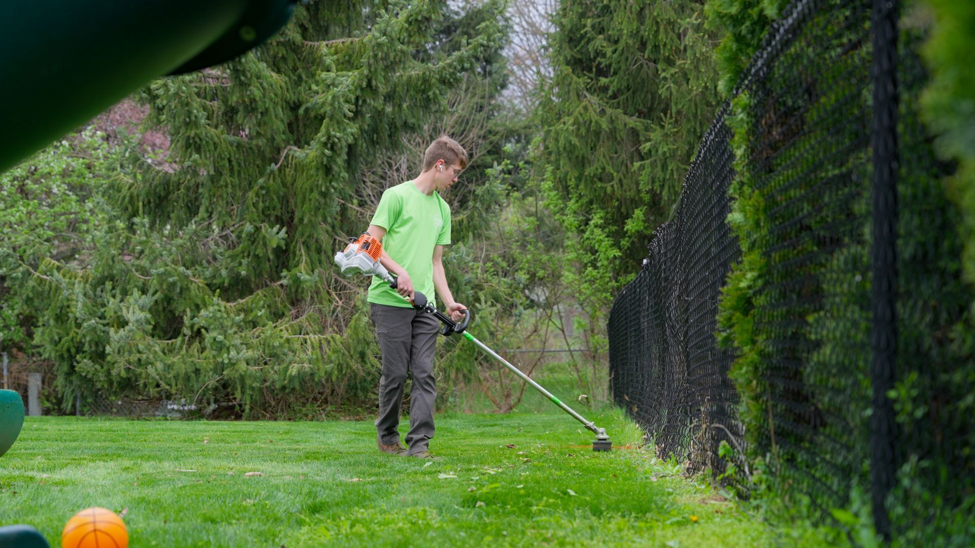 Person using a string trimmer in a grassy backyard near a black fence and trees