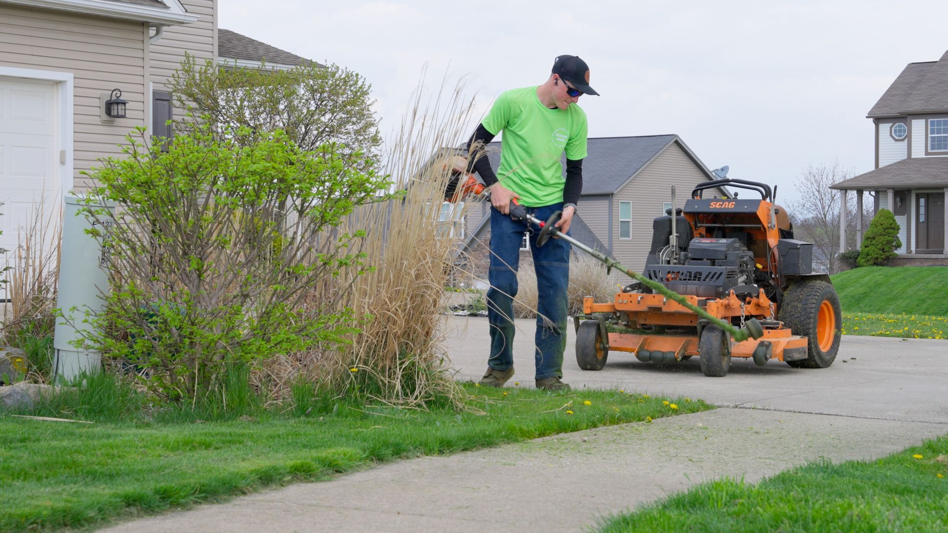 Person mowing a suburban lawn with a push mower beside a sidewalk and houses