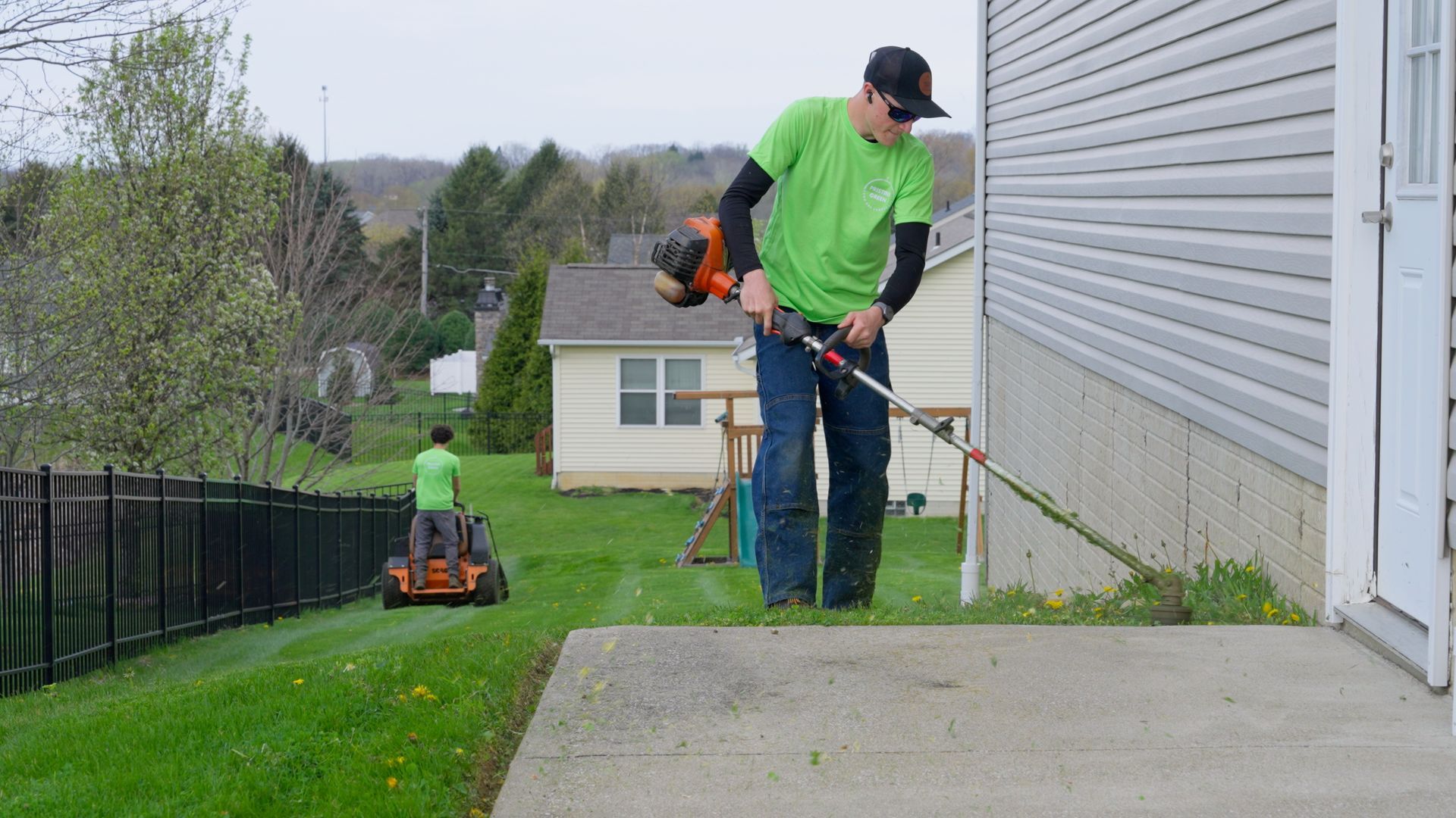 Workers using trimmer and mower along a house and fence in a grassy yard