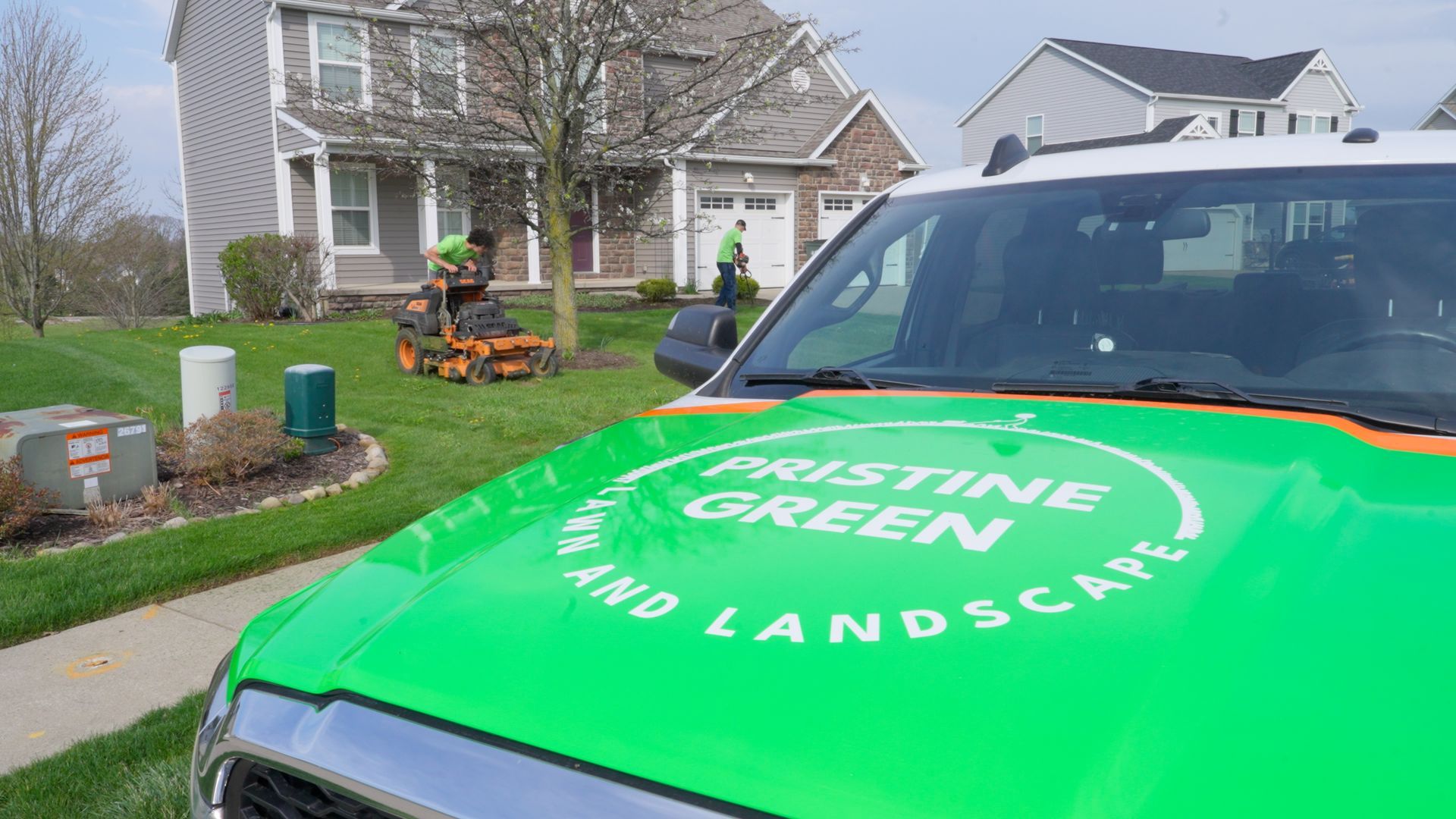 Green lawn care truck in a suburban yard with a worker mowing the grass.