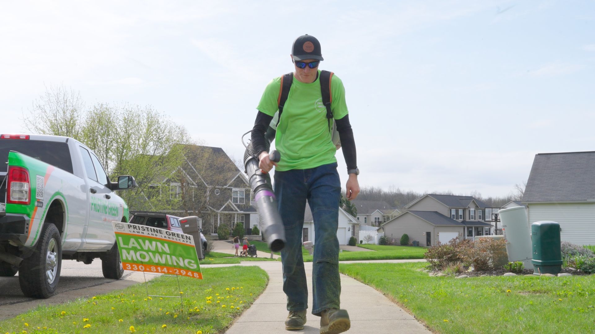 Man in green shirt and backpack walking a sidewalk while carrying a backpack sprayer in a suburban neighborhood