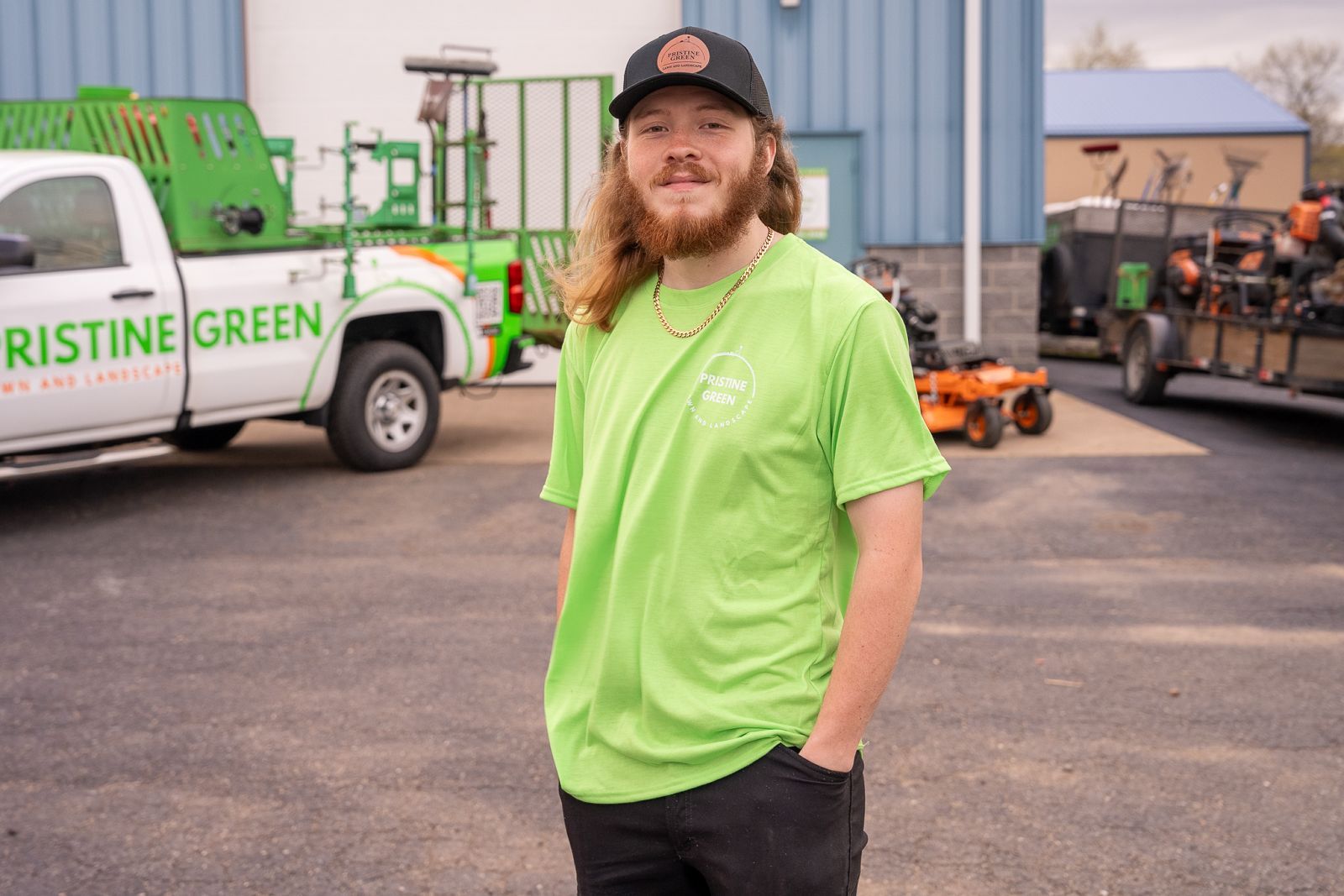 Man in lime green shirt and black cap stands in a parking lot with work trucks and equipment behind him