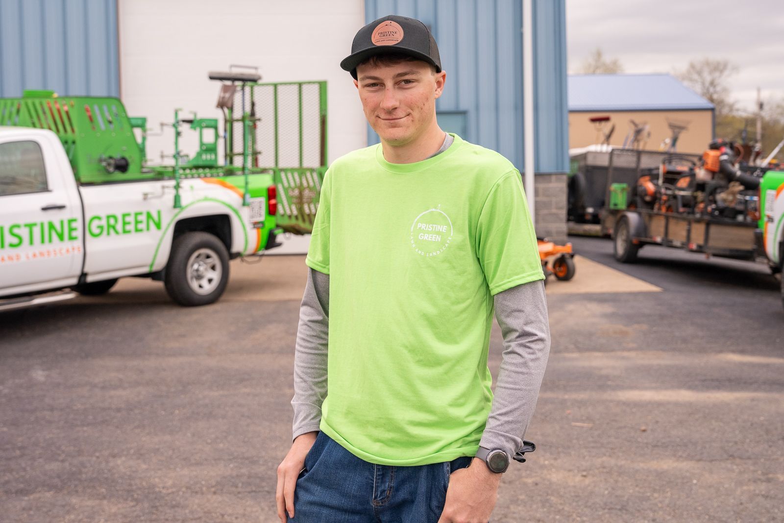 Young man in a green shirt and cap standing in a parking lot with work trucks and equipment behind him