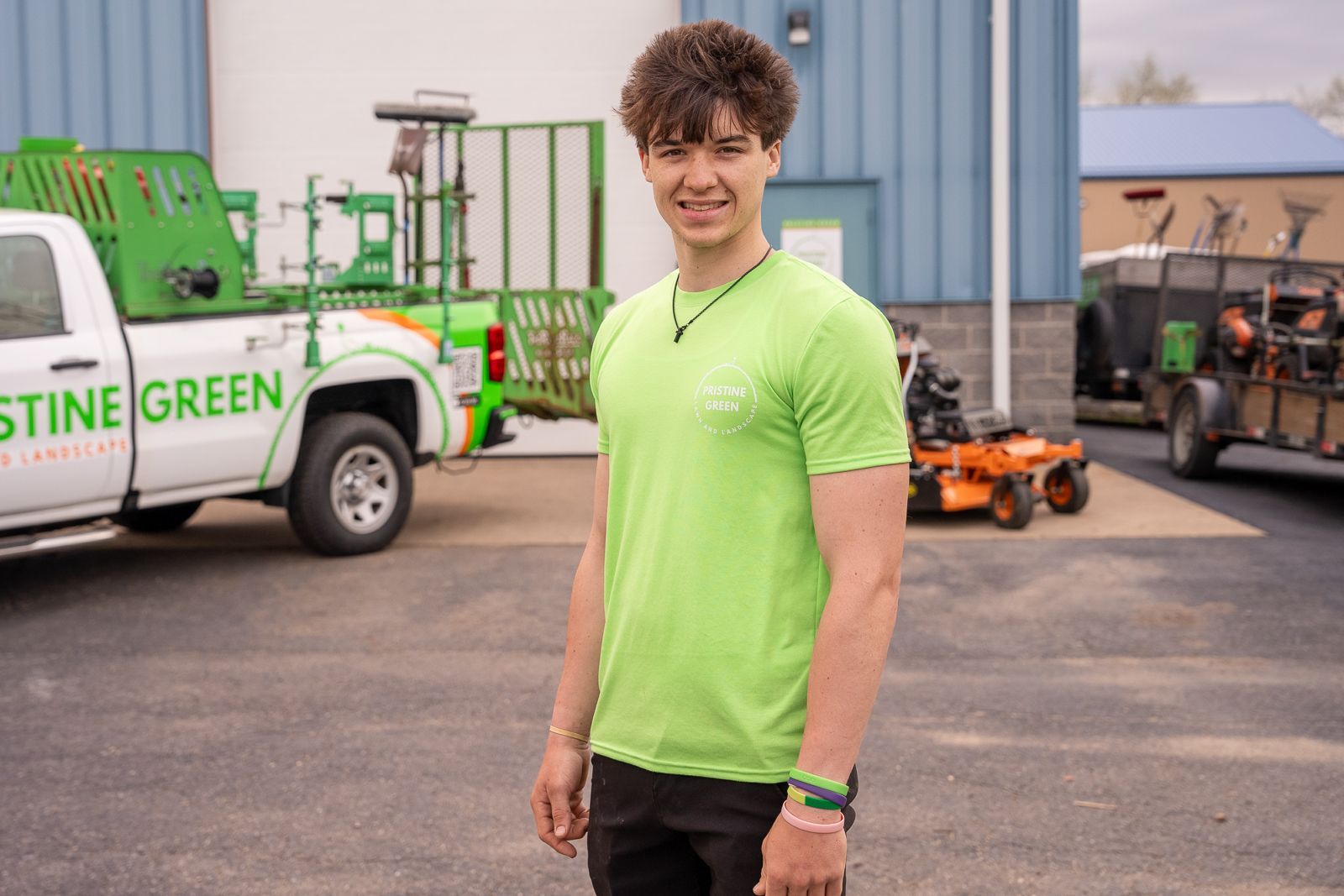 Person in a neon green shirt standing in a parking lot beside landscaping trucks and equipment