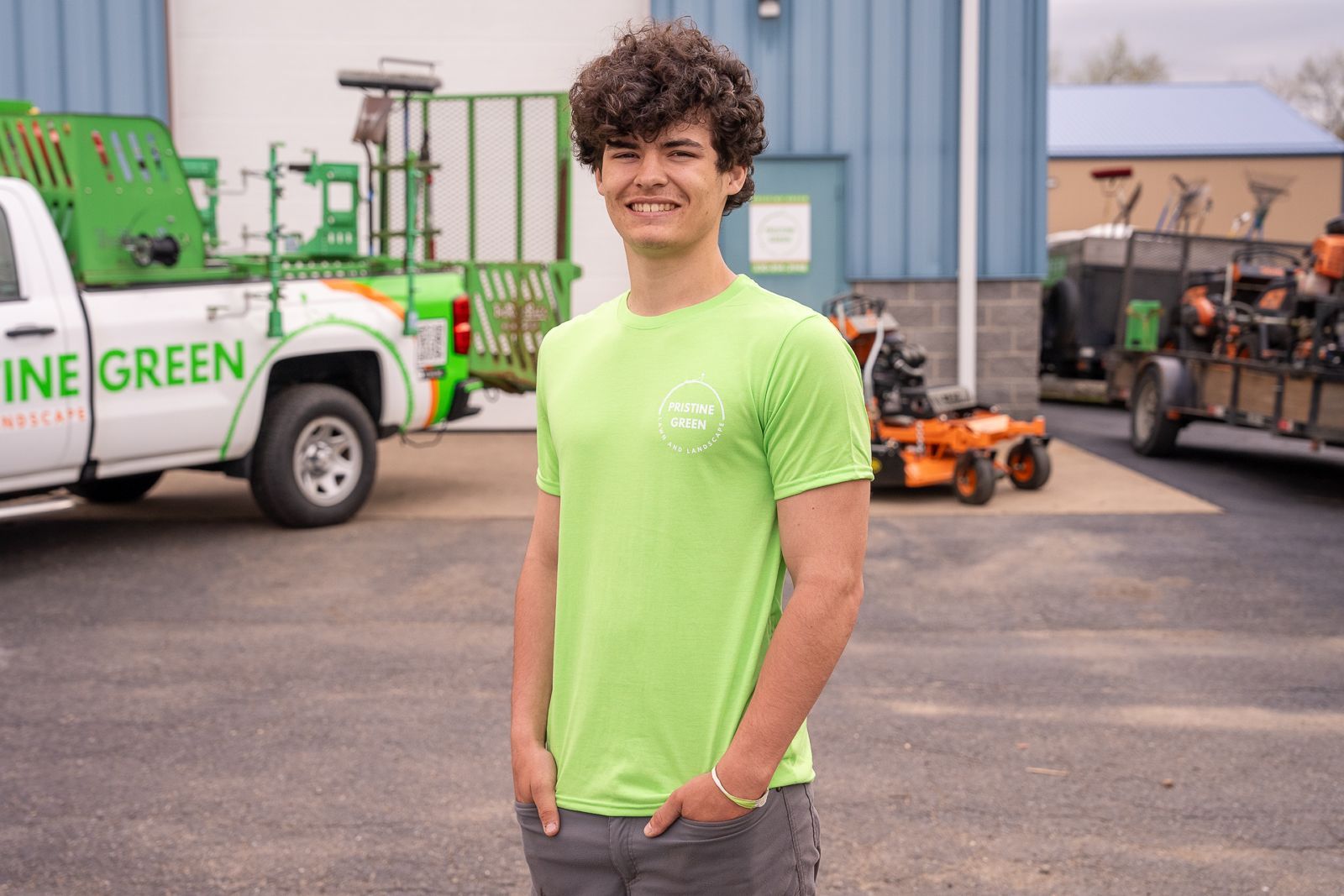 Man in neon green shirt standing in an industrial lot with utility vehicles and equipment behind him
