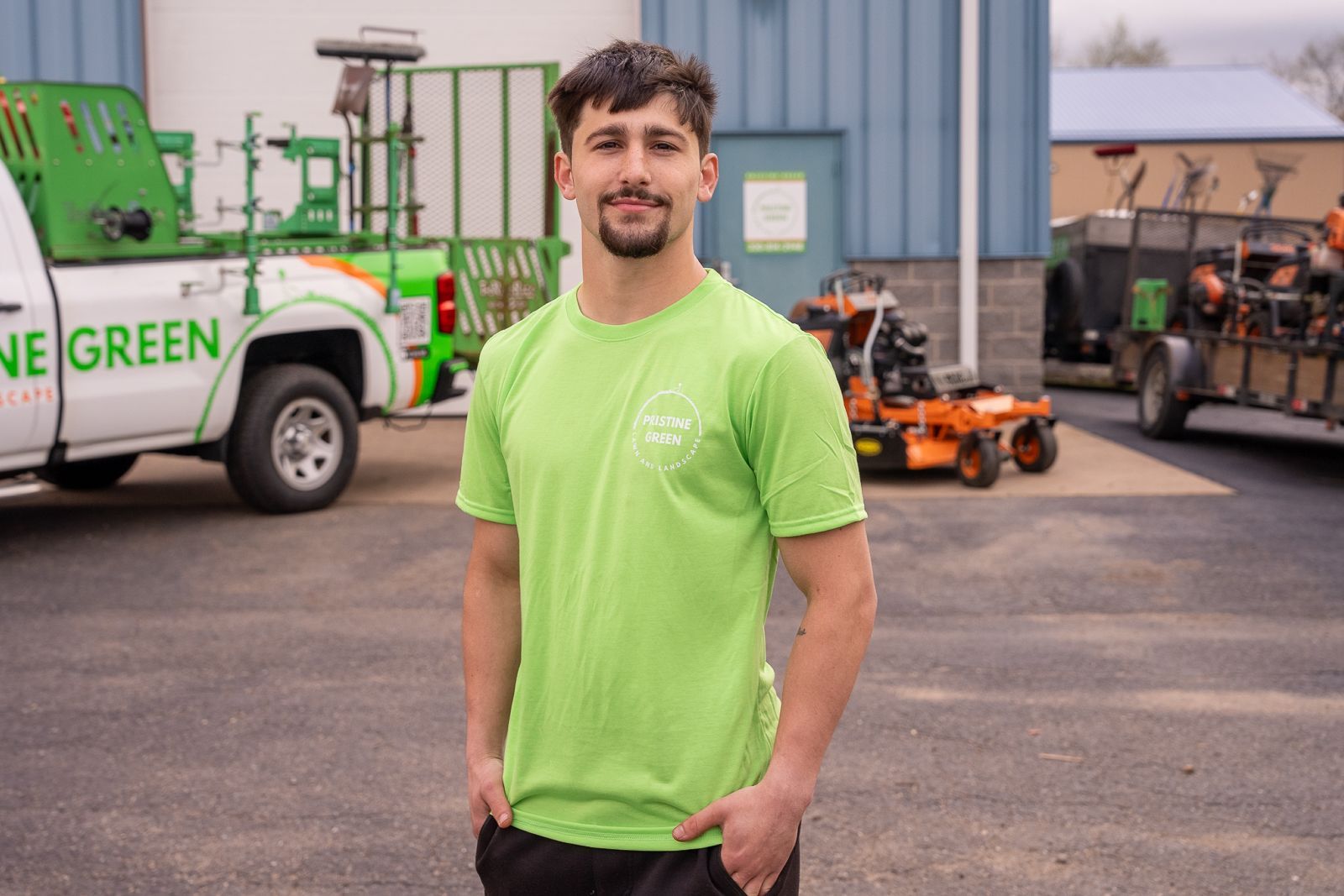 Man in a bright green shirt standing in a work yard with utility vehicles and equipment in the background