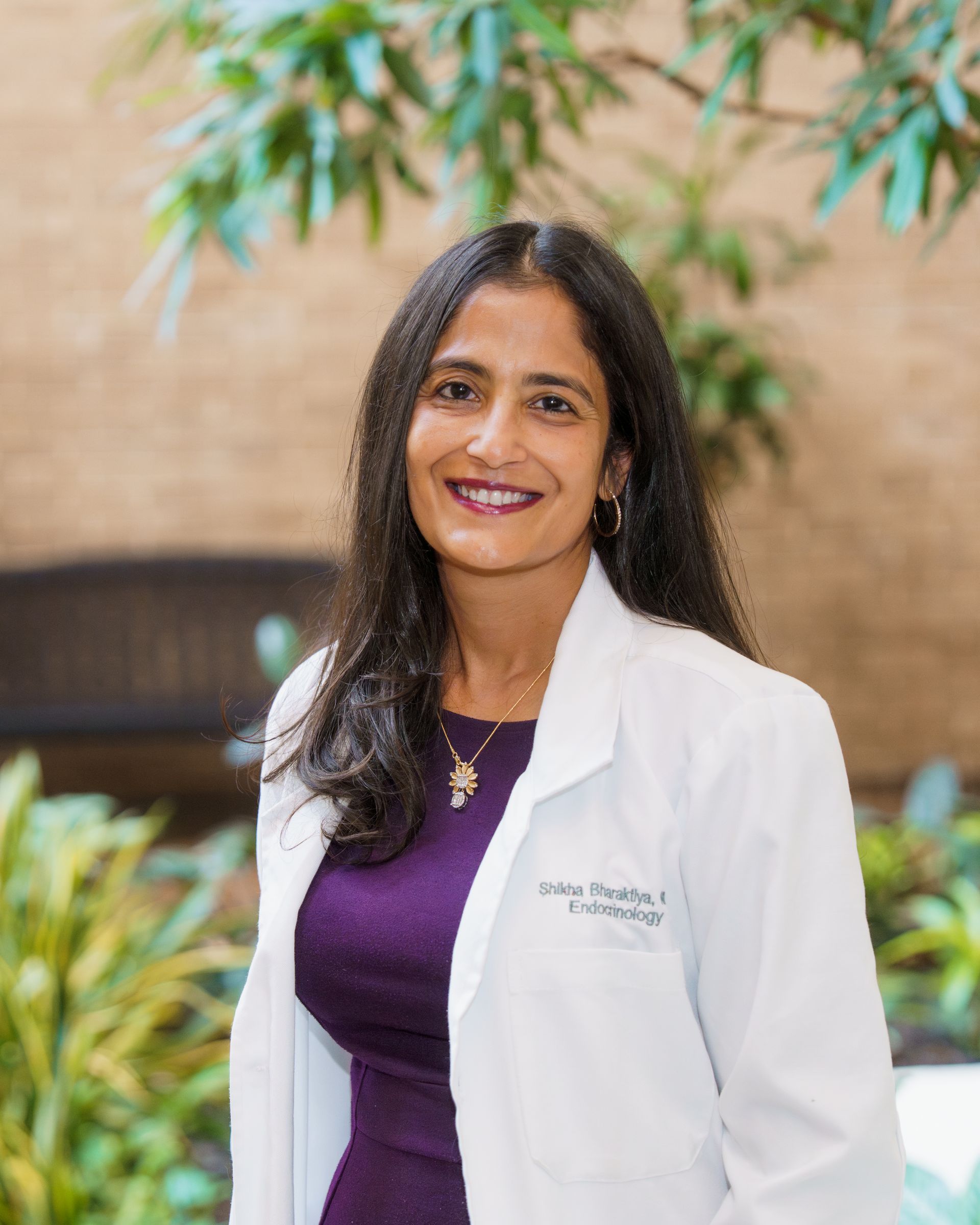 Smiling medical professional in a white coat and purple top, standing outdoors with greenery behind her