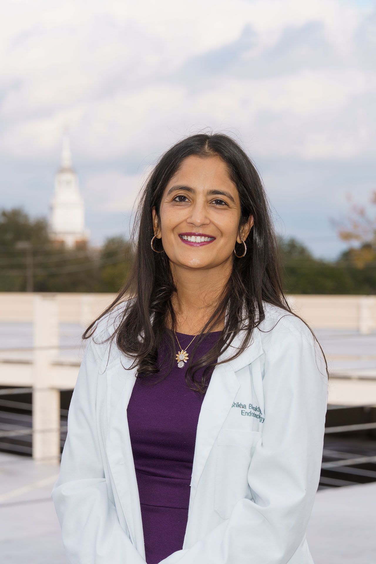 Woman in a white lab coat and purple top smiling outdoors with a blurred building in the background