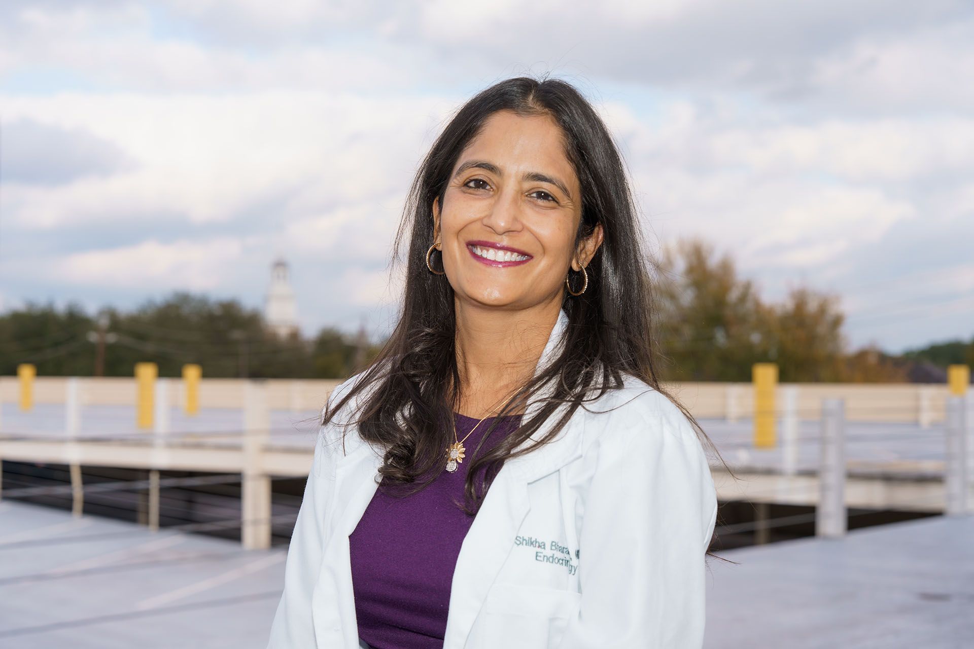 Smiling person in a white coat and purple top standing outdoors on a rooftop deck