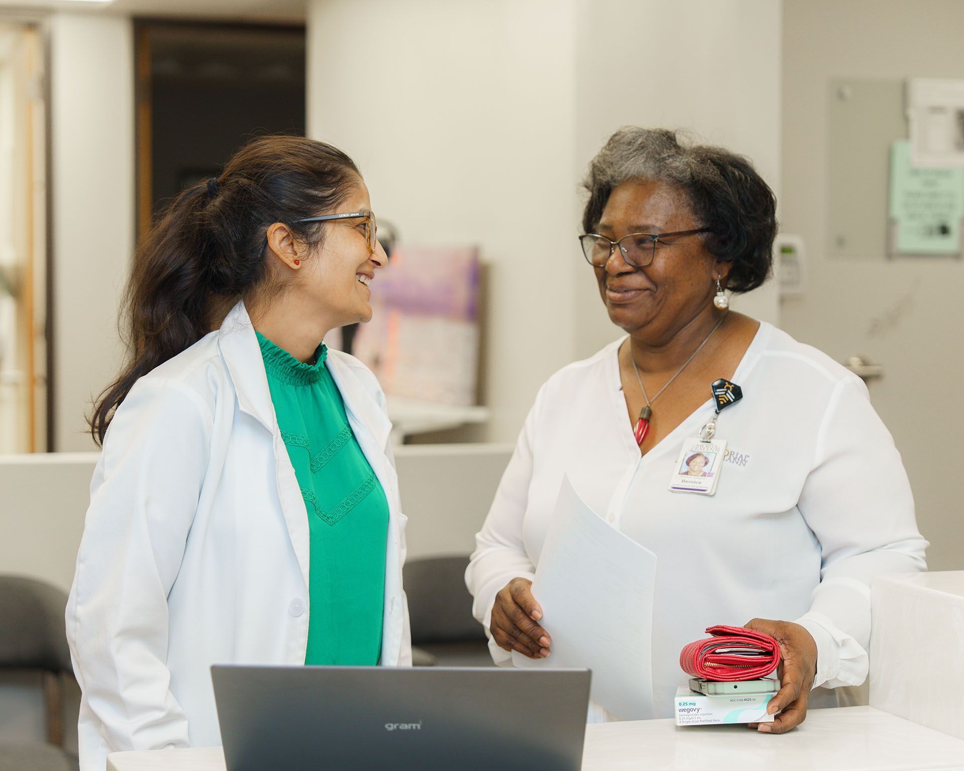 Two medical staff in white coats talking at a hospital counter, one holding papers and smiling.