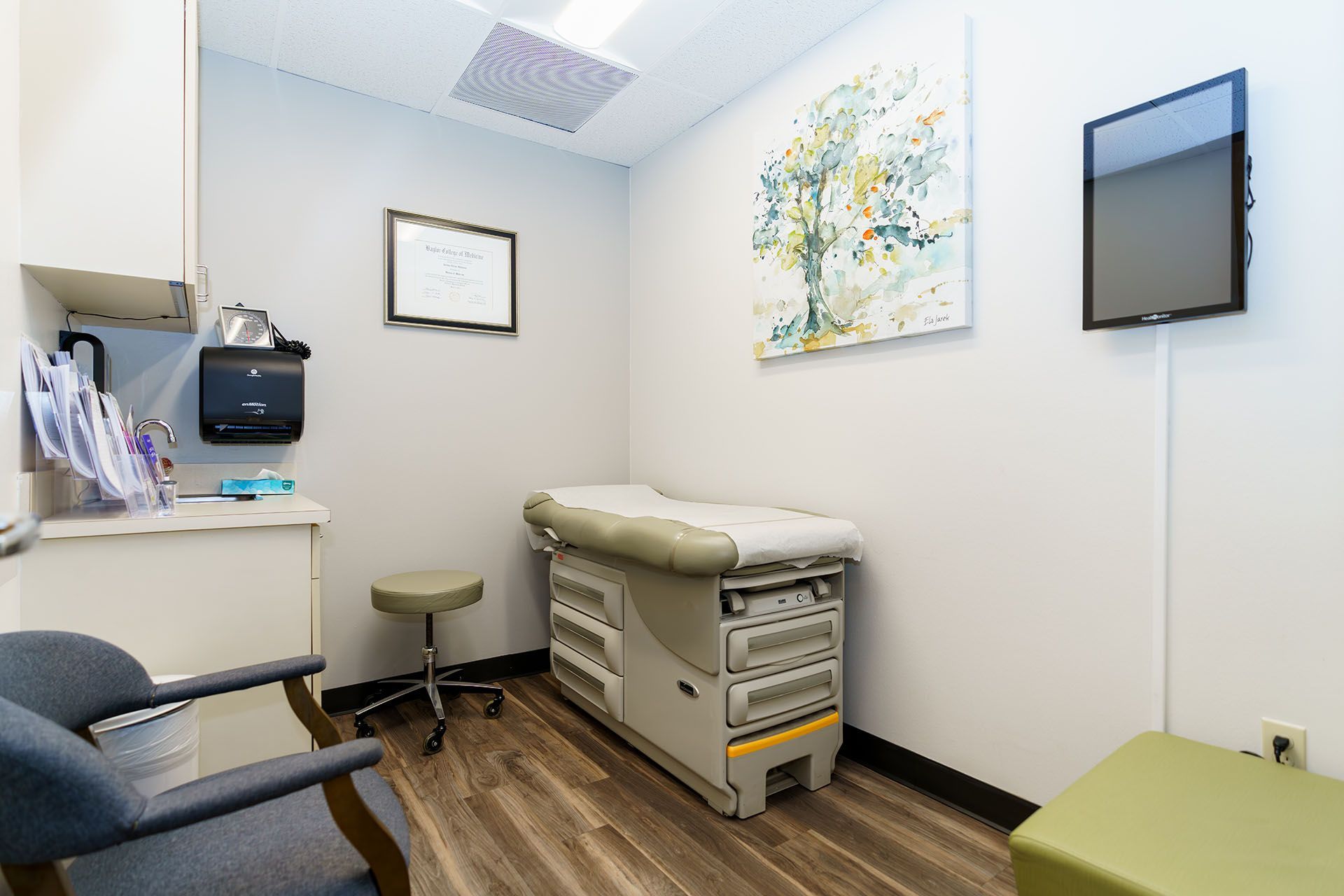 Small medical exam room with exam table, chair, wall monitor, and artwork on white walls