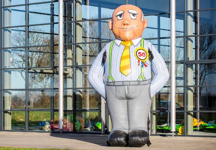 Opblaasbare figuur van een man met bretels, een stropdas en een 50-badge, staand voor een gebouw.