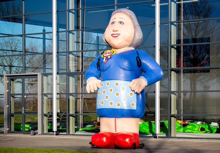 Een groot standbeeld van een vrouw in een blauwe outfit, rode schoenen en een schort staat voor een glazen gebouw.