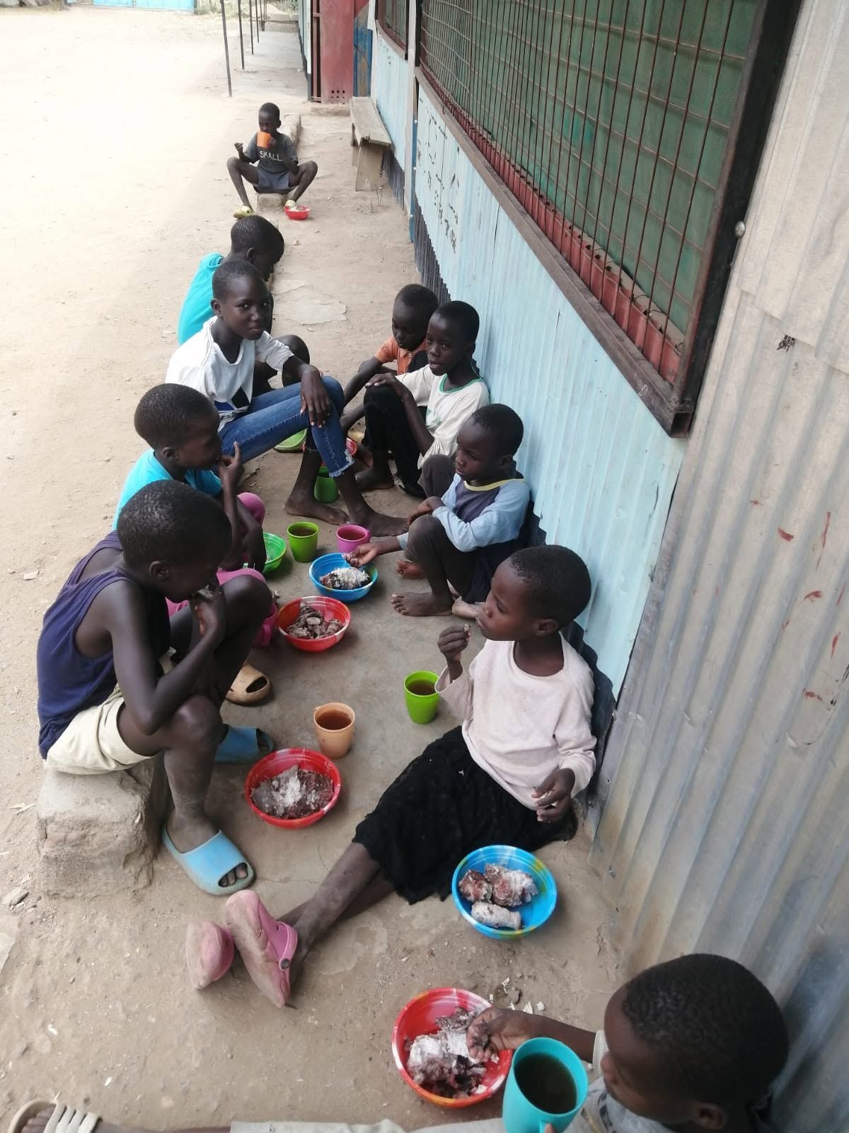 Children eating meals in a line along a building wall, with colorful plates on the ground.