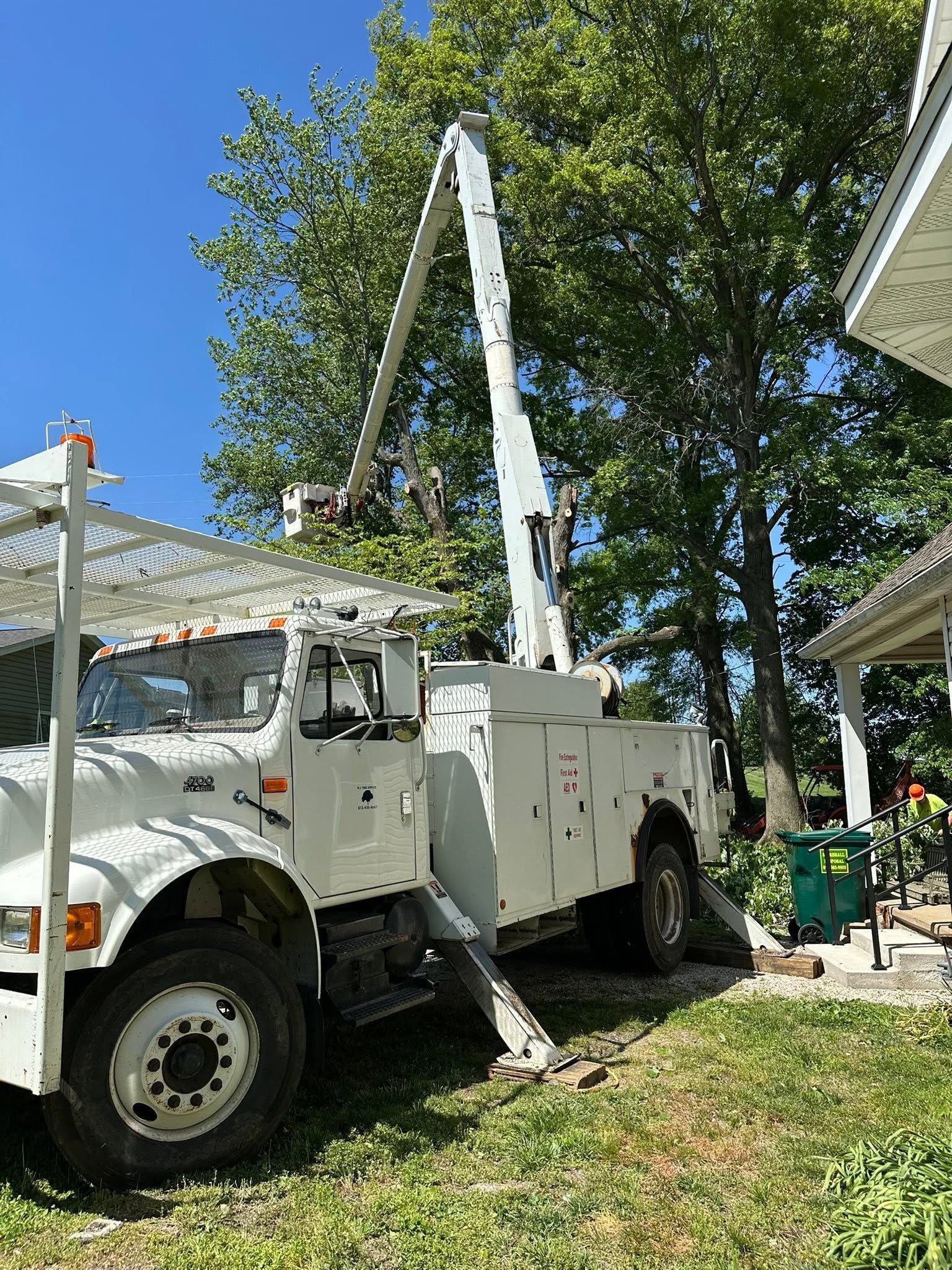 White utility truck with raised boom parked in a leafy yard beside a house.