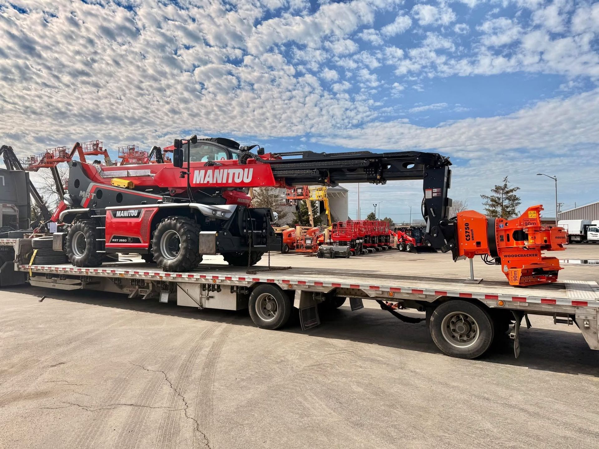 Red Manitou telehandler on a flatbed trailer in a lot under a cloudy sky
