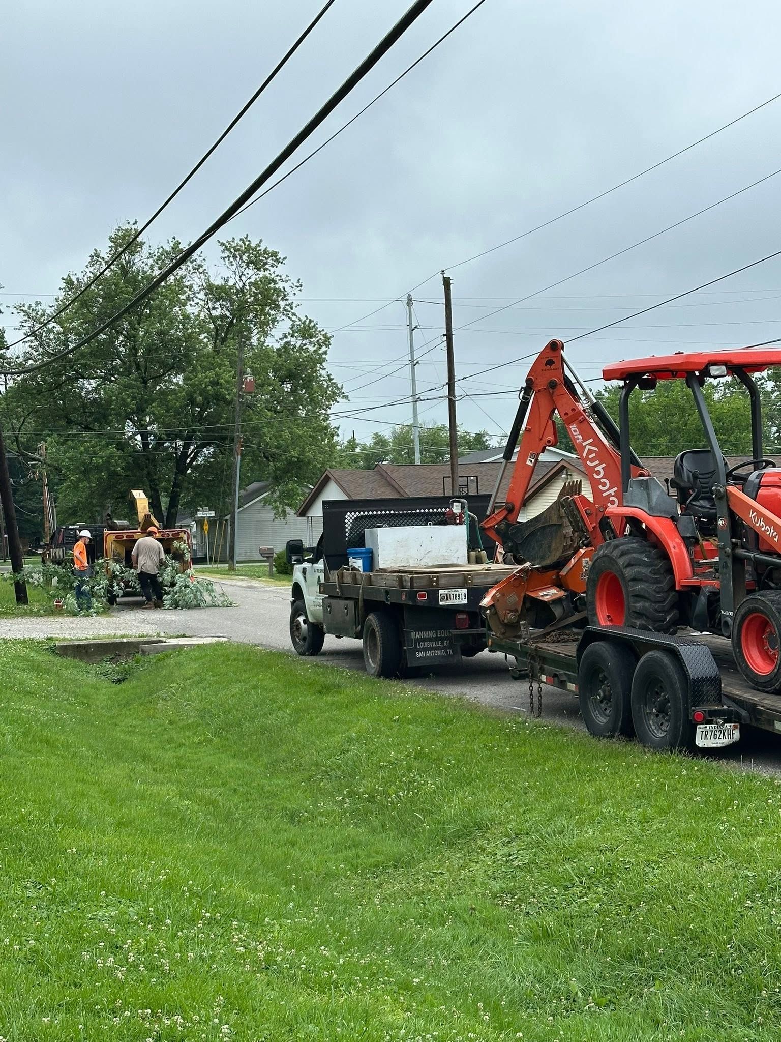 Roadside utility crew with backhoe and trailer working near power lines on a wet street