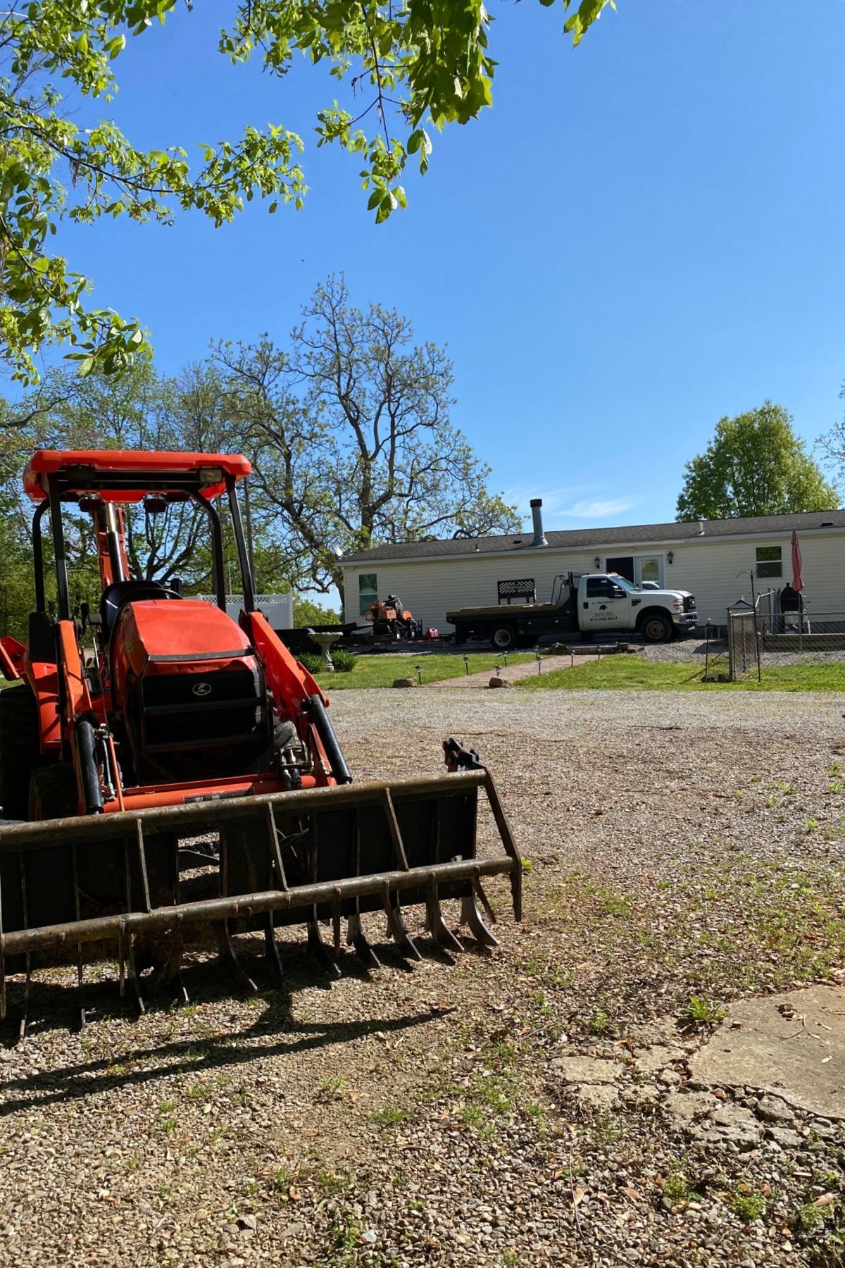 Orange tractor with front loader in a gravel yard near a house and parked truck under a blue sky