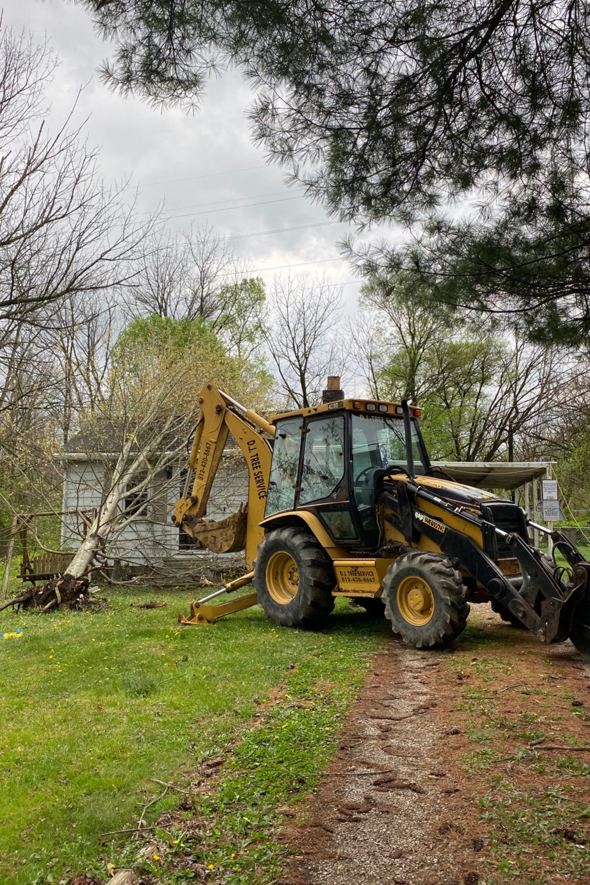 Yellow backhoe parked on a grassy path beside a house and trees on an overcast day