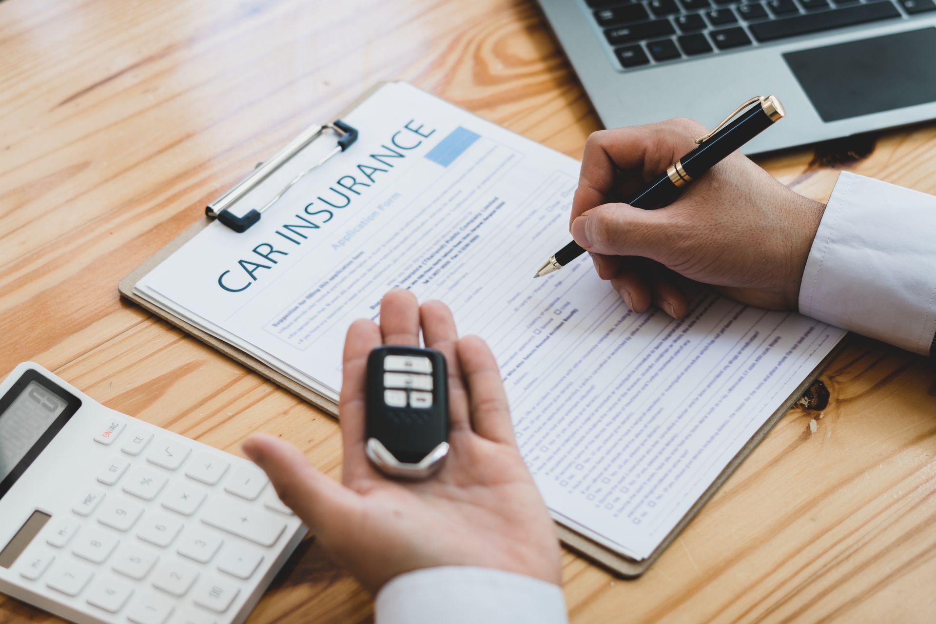 Man signing car insurance form with pen, holding car key, calculator and paperwork on desk.